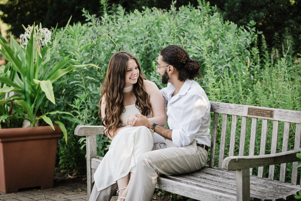 Engaged couple in coordinated cream outfits seated on garden bench at Deep Cut Gardens