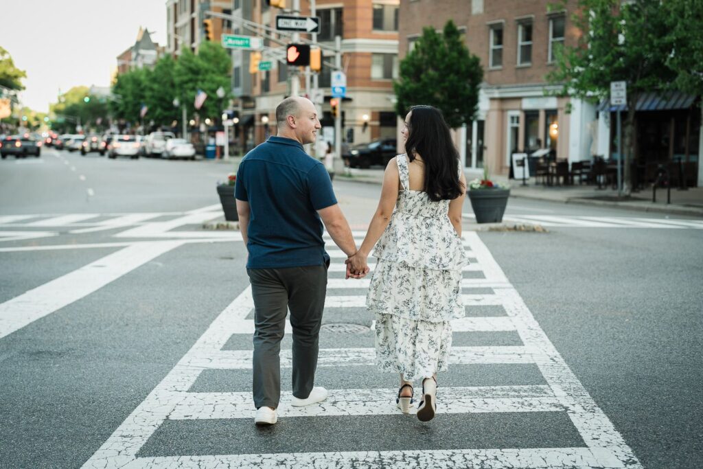 Engaged couple crossing a downtown Morristown street hand in hand and looking at each other