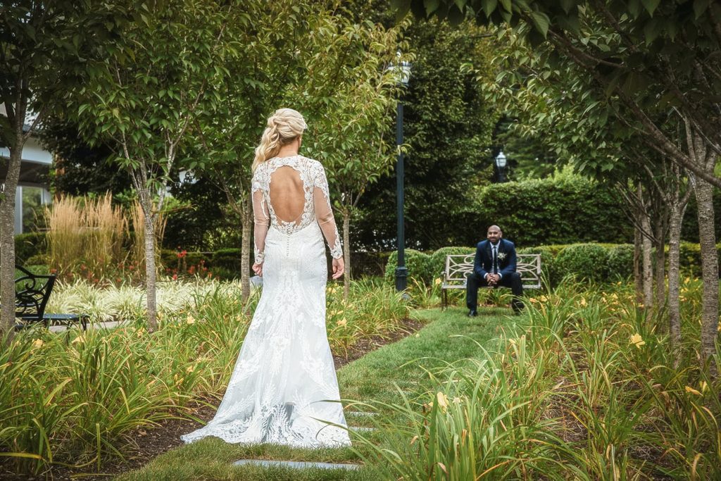 Bride walking a garden path toward her seated groom for a first look at The Rockleigh