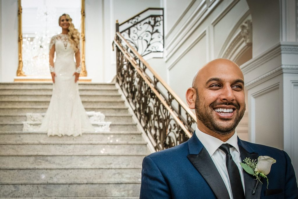 Groom laughing during a first look as his bride waits on the marble staircase at The Rockleigh