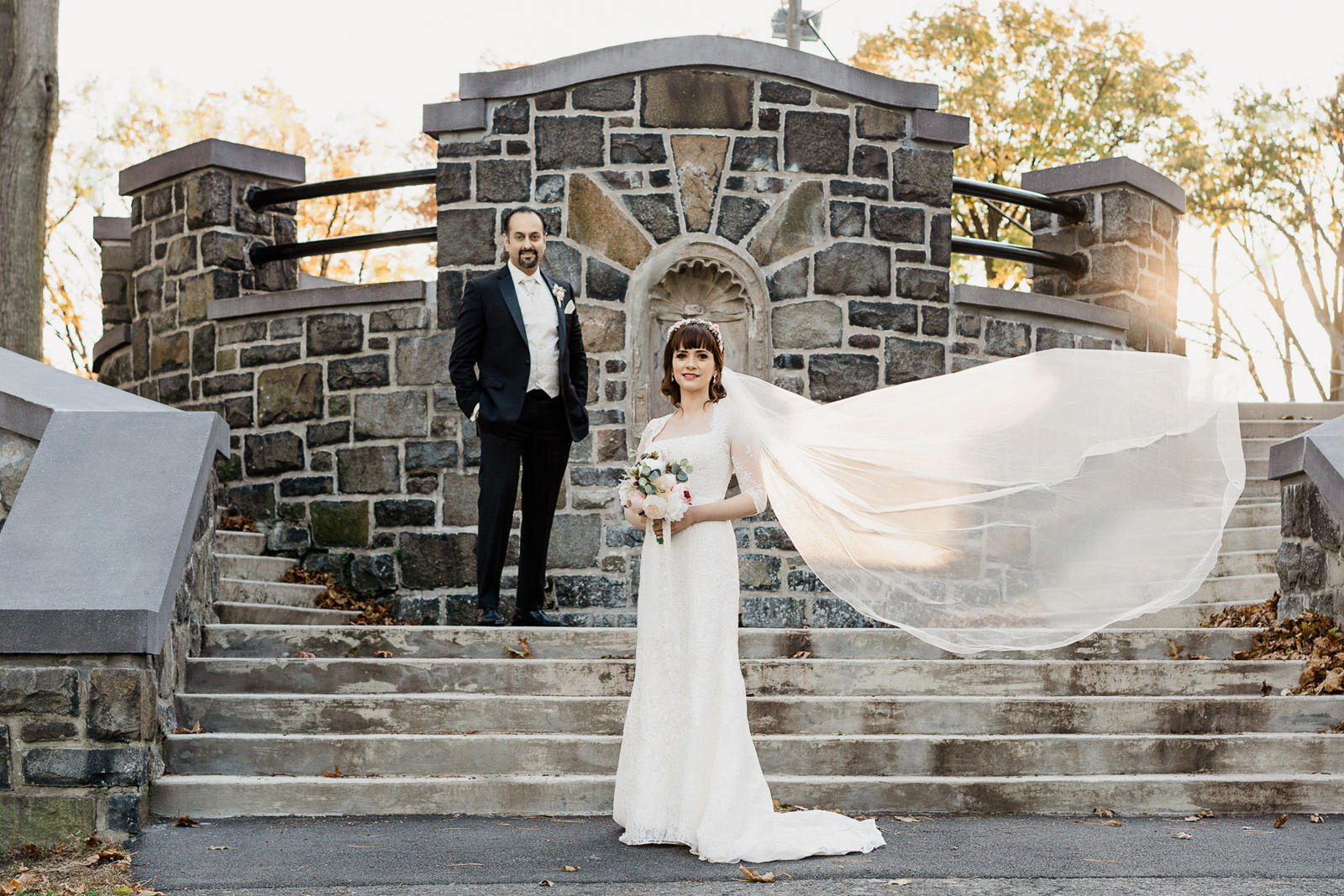 Bride and groom on stone staircase with veil glowing in late-day light