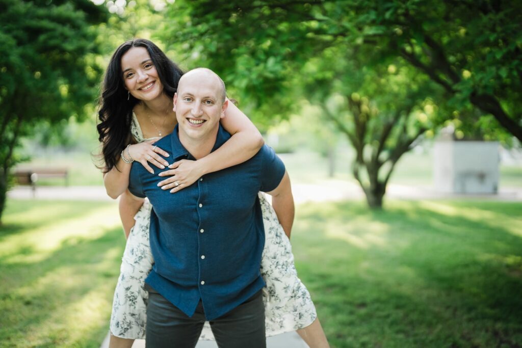 Engaged couple smiling during a piggyback ride under the trees at Burnham Park Morristown