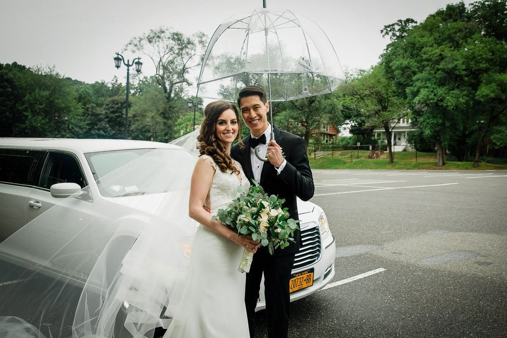 Bride and groom under a clear umbrella beside a wedding limousine on a rainy day