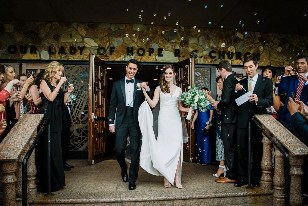 Newlyweds exiting a church under a covered entrance as guests blow bubbles