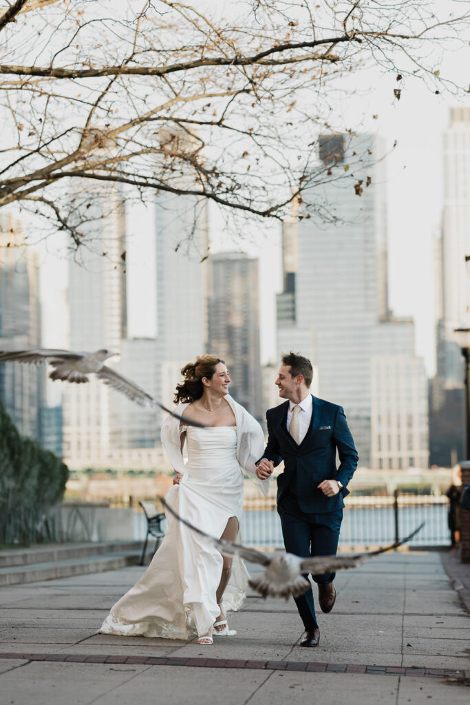 Bride and groom running along a waterfront with city skyline and birds behind them