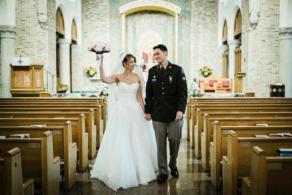 Bride raising her bouquet beside the groom in the empty aisle of a Harrison NY church