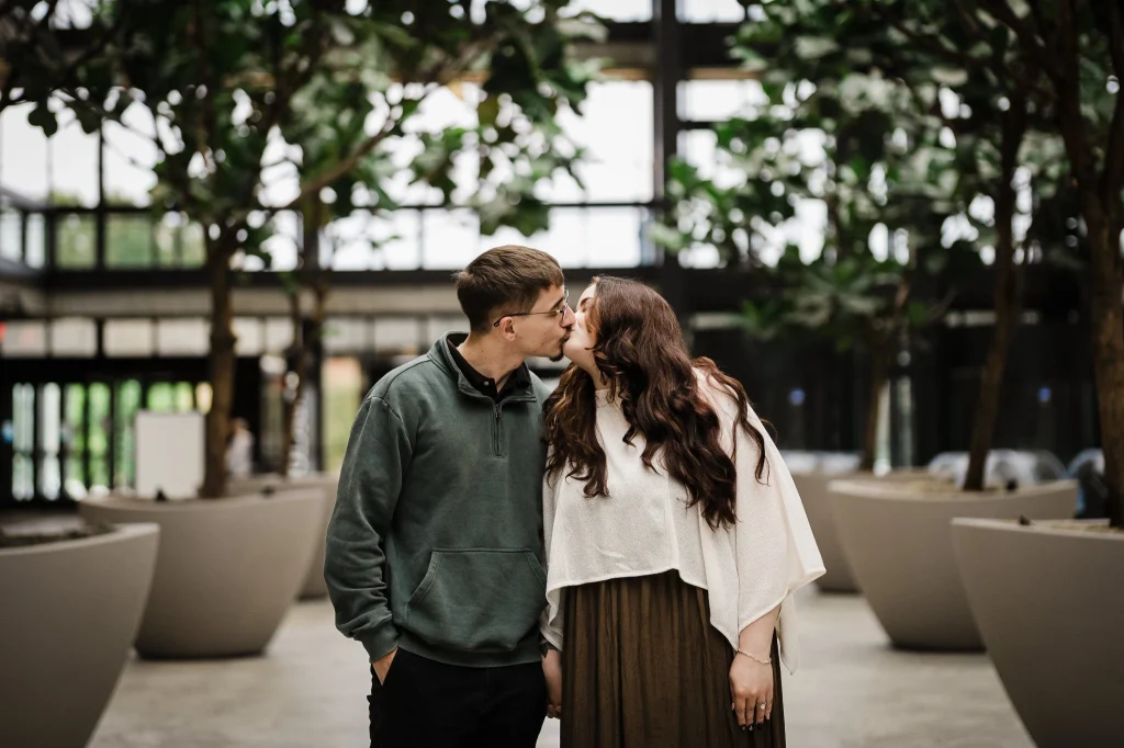 Engaged couple sharing a kiss among large planters and indoor trees at Bell Works