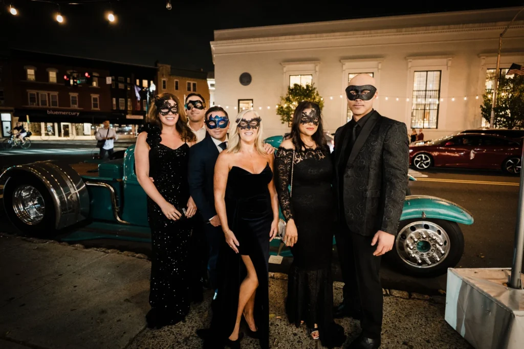 Six masked guests in black formalwear pose beside a hot rod outside James Ward Mansion